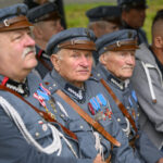Grupa starszych mężczyzn w szarych mundurach z medalami siedzi w rzędzie. Mundury ozdobione są kolorowymi odznaczeniami. W tle rozmyte otoczenie natury.