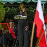 Na scenie pod zielonym namiotem stoi kobieta w czarnym stroju, trzyma mikrofon. Obok muzycy w czerwonych mundurach grają na instrumentach. W tle widać polskie flagi.