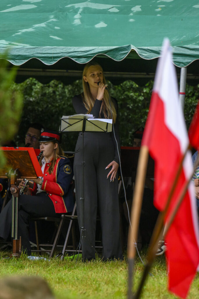 Na scenie pod zielonym namiotem stoi kobieta w czarnym stroju, trzyma mikrofon. Obok muzycy w czerwonych mundurach grają na instrumentach. W tle widać polskie flagi.