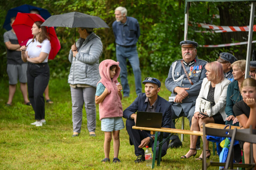 Grupa ludzi na zewnątrz podczas wydarzenia. Niektórzy siedzą, inni stoją pod parasolami. Tło to zielone drzewa i trawa.