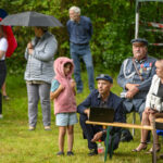 Grupa ludzi na zewnątrz podczas wydarzenia. Niektórzy siedzą, inni stoją pod parasolami. Tło to zielone drzewa i trawa.