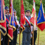 Grupa osób w mundurach stoi z kolorowymi sztandarami. Dominują barwy czerwone, niebieskie i białe. Tło to zielone drzewa.