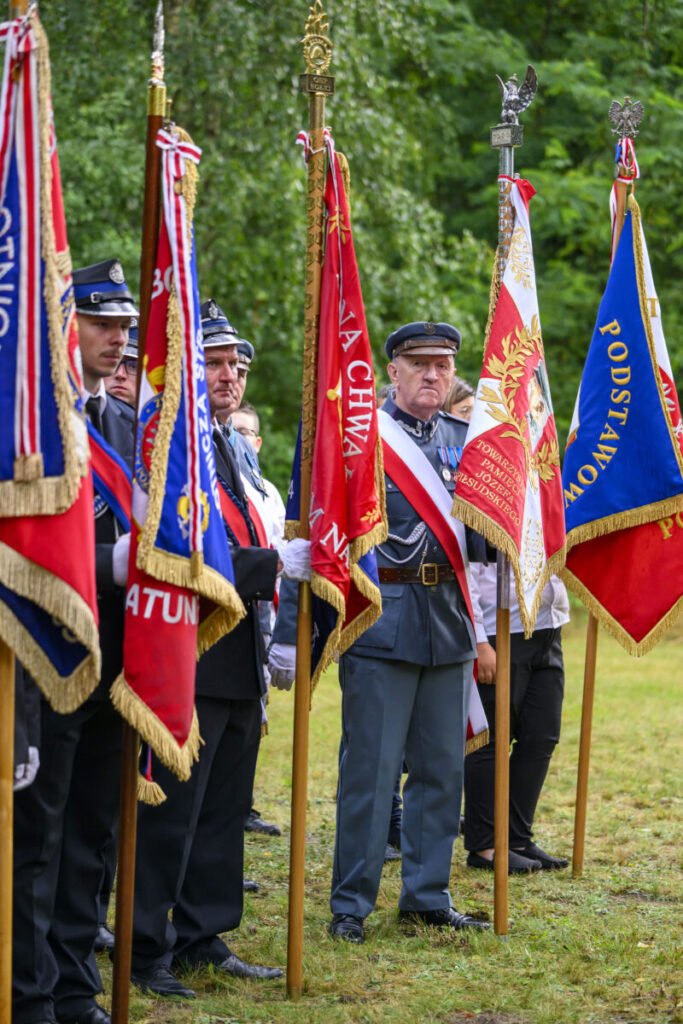 Grupa osób w mundurach stoi z kolorowymi sztandarami. Dominują barwy czerwone, niebieskie i białe. Tło to zielone drzewa.