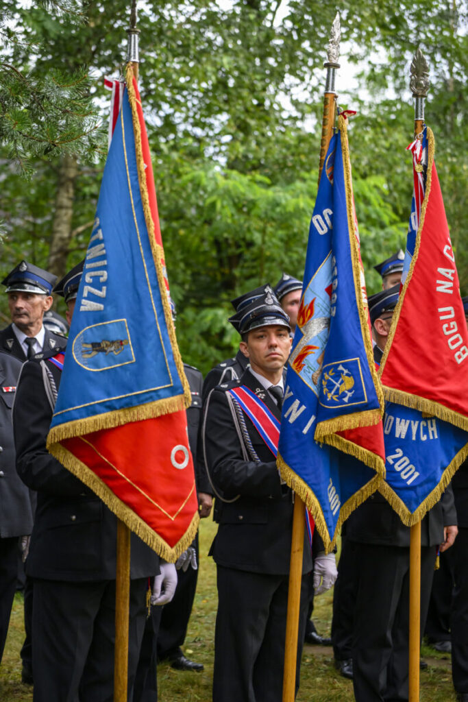 Strażacy w czarnych mundurach stoją z kolorowymi sztandarami: niebieskim i czerwonym. W tle zielone drzewa.