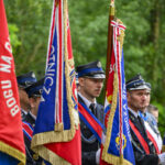 Strażacy w mundurach stoją z flagami w barwach czerwonych i niebieskich. Tło stanowią zielone drzewa. Uroczysty charakter zdjęcia.