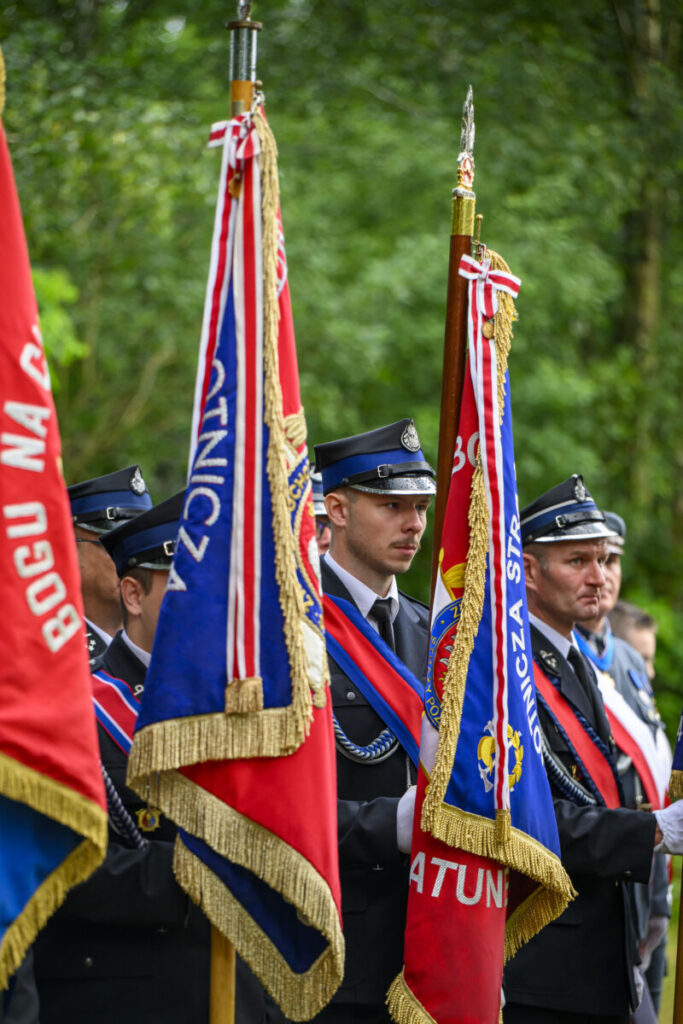 Strażacy w mundurach stoją z flagami w barwach czerwonych i niebieskich. Tło stanowią zielone drzewa. Uroczysty charakter zdjęcia.