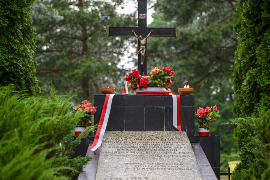 Czarny krzyż z Jezusem na tle zielonych drzew. Czerwone kwiaty i biało-czerwone wstęgi ozdabiają pomnik. Wokół płoną świece.