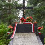 Czarny krzyż z Jezusem na tle zielonych drzew. Czerwone kwiaty i biało-czerwone wstęgi ozdabiają pomnik. Wokół płoną świece.