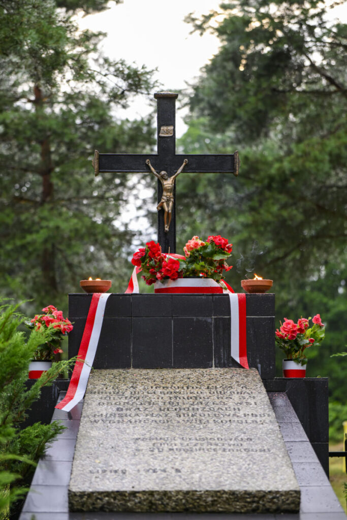 Czarny krzyż z figurą Jezusa na tle zielonych drzew. Przed krzyżem czerwone kwiaty i biało-czerwone wstążki. Przy krzyżu płoną znicze.