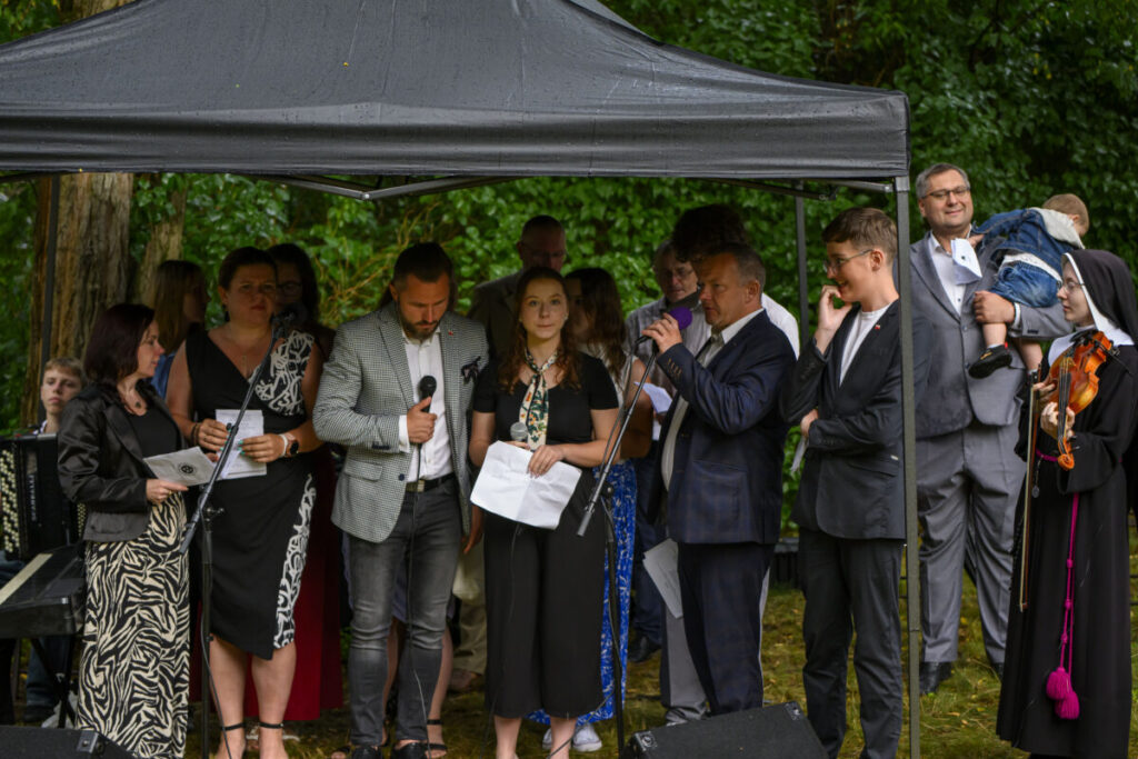 Grupa ludzi pod czarnym namiotem, trzymają mikrofony i kartki. W tle zielone drzewa. Uczestnicy w eleganckich strojach.