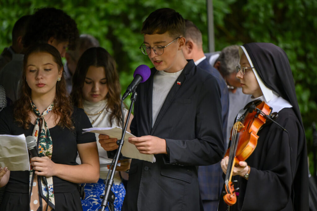 Grupa młodych ludzi odczytuje teksty na zewnątrz, na tle zieleni. Ubrani elegancko; jedna osoba trzyma skrzypce. Mikrofon ma fioletową osłonę.