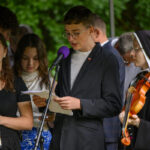 Grupa młodych ludzi odczytuje teksty na zewnątrz, na tle zieleni. Ubrani elegancko; jedna osoba trzyma skrzypce. Mikrofon ma fioletową osłonę.