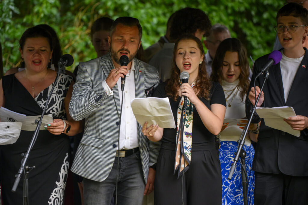 Grupa osób śpiewa na scenie z mikrofonami. Tło z zielonymi liśćmi. Ubrani są elegancko, w ubrania w różnych kolorach.