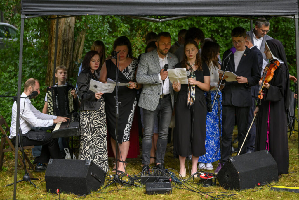 Grupa osób śpiewa pod zadaszeniem, obok pianista w białej koszuli. Tło to zieleń drzew, a na ziemi leżą przewody.