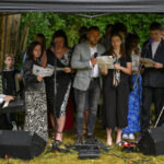 Grupa osób śpiewa pod zadaszeniem, obok pianista w białej koszuli. Tło to zieleń drzew, a na ziemi leżą przewody.