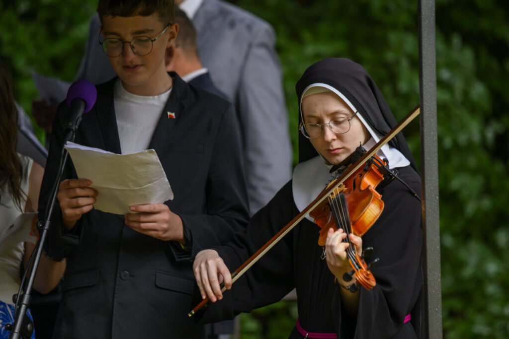 Na zdjęciu stoi osoba w zakonnym habicie grająca na skrzypcach i mężczyzna w ciemnej marynarce. Tło jest zielone i zamglone. Obok nich znajduje się mikrofon.