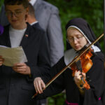 Na zdjęciu stoi osoba w zakonnym habicie grająca na skrzypcach i mężczyzna w ciemnej marynarce. Tło jest zielone i zamglone. Obok nich znajduje się mikrofon.