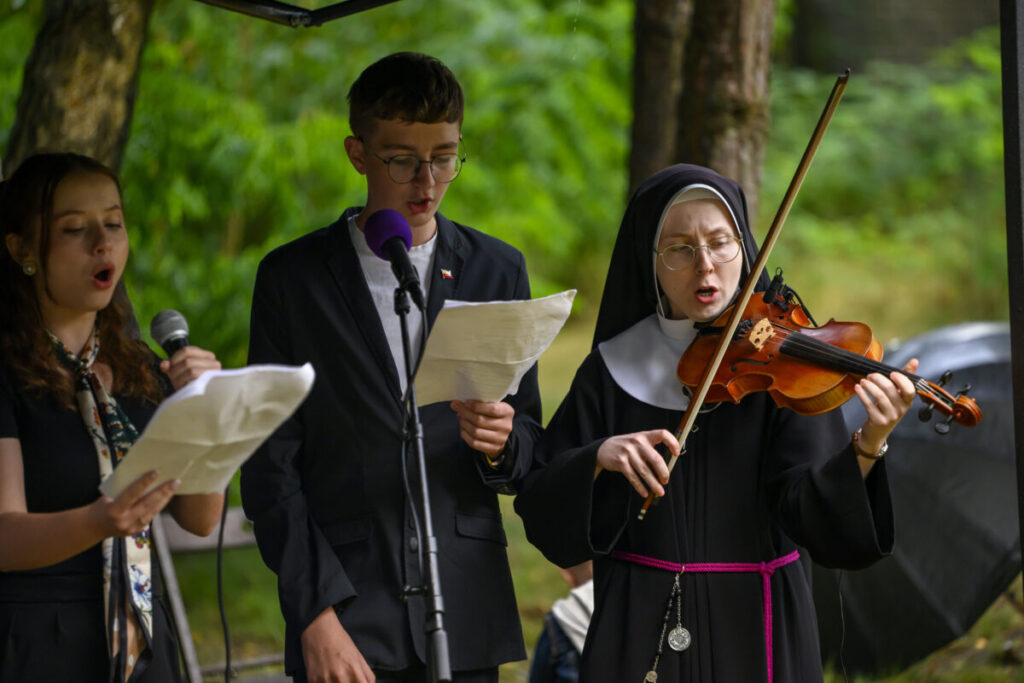 Trzy osoby śpiewają i grają na instrumentach na tle zieleni. Kobieta w habicie gra na skrzypcach. Mężczyzna i kobieta trzymają mikrofony.