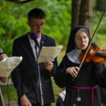 Trzy osoby śpiewają i grają na instrumentach na tle zieleni. Kobieta w habicie gra na skrzypcach. Mężczyzna i kobieta trzymają mikrofony.