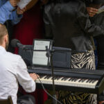 Na zdjęciu mężczyzna w białej koszuli gra na pianinie. Przed nim nuty na tablet; wokół grupa ludzi z kartkami. Tło ciemne, kolorowe ubrania.