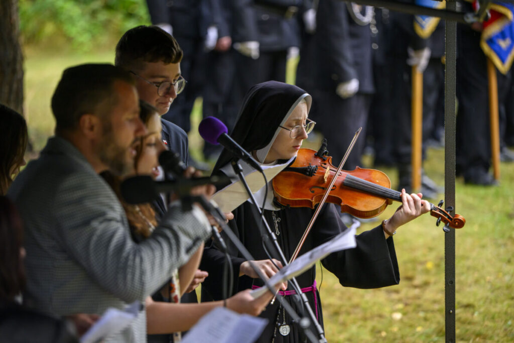 Na zdjęciu widać zakonnicę grającą na skrzypcach, obok grupy osób z mikrofonami. Tło jest zielone, przypominające park. Ubrania są ciemne.