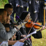 Na zdjęciu widać zakonnicę grającą na skrzypcach, obok grupy osób z mikrofonami. Tło jest zielone, przypominające park. Ubrania są ciemne.