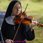 Zakonnica w czarnym habicie gra na skrzypcach. Tło jest rozmyte, zielone. Wokół niej jest spokojna atmosfera.