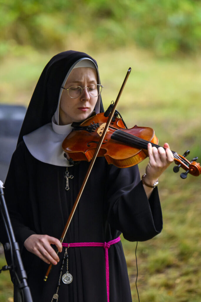 Zakonnica w czarnym habicie gra na skrzypcach. Tło jest rozmyte, zielone. Wokół niej jest spokojna atmosfera.