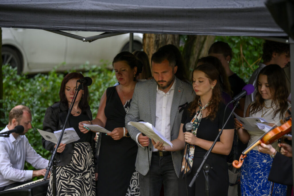 Grupa osób śpiewa, trzymając nuty. Tło to zieleń, za nimi samochód. Jedna osoba gra na pianinie, mikrofony z przodu.