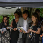 Grupa osób śpiewa, trzymając nuty. Tło to zieleń, za nimi samochód. Jedna osoba gra na pianinie, mikrofony z przodu.