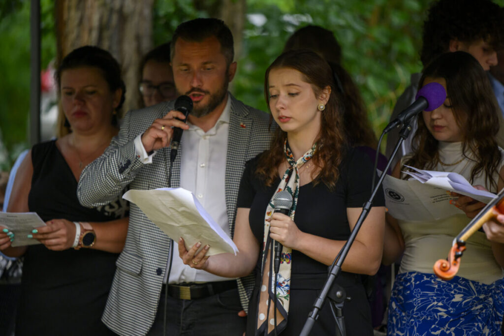 Grupa osób śpiewa z mikrofonami, trzymając kartki. Na pierwszym planie mężczyzna i kobieta. Tło rozmyte, zielone drzewa.
