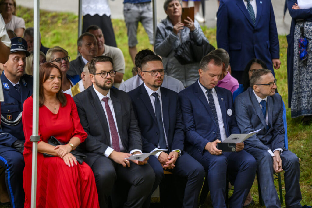 Grupa osób siedzących na zewnątrz na ławkach, ubranych elegancko. Kobieta w czerwonej sukni, mężczyźni w garniturach. W tle trawa i ludzie stojący.