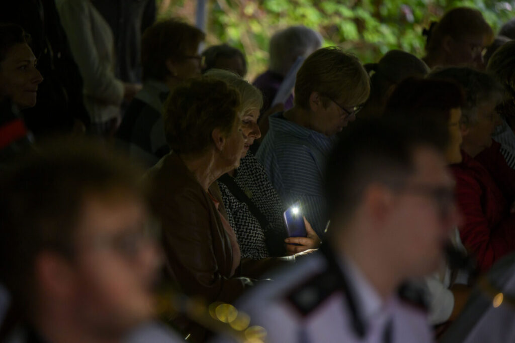 Grupa osób siedzących na widowni, jedna osoba trzyma świecący telefon. Tło jest ciemne, nieostre, z zielenią. Jasne światło skupia się na twarzy kobiety.