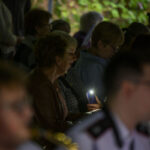 Grupa osób siedzących na widowni, jedna osoba trzyma świecący telefon. Tło jest ciemne, nieostre, z zielenią. Jasne światło skupia się na twarzy kobiety.