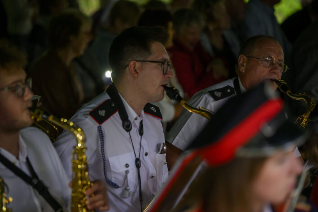 Orkiestra dęta gra koncert, muzycy w białych mundurach i czapkach; mosiężne instrumenty błyszczą na tle tłumu.