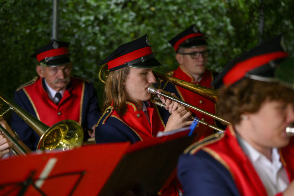 Osoby w czerwonych mundurach i czapkach grają na trąbkach. Tło zielone, przypomina drzewa. Wyrazistość kolorów podkreśla uroczystość.
