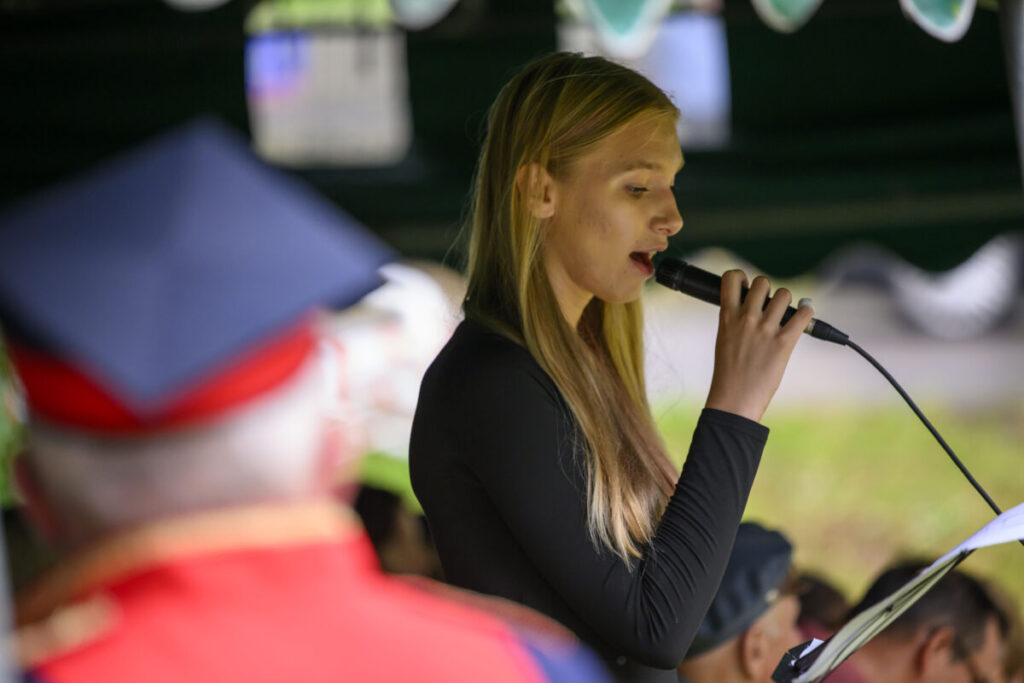 Młoda kobieta w czarnej bluzce śpiewa do mikrofonu. W tle rozmyte postacie i zielone otoczenie. Z przodu osoba w czerwonym stroju.