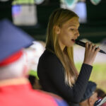 Młoda kobieta w czarnej bluzce śpiewa do mikrofonu. W tle rozmyte postacie i zielone otoczenie. Z przodu osoba w czerwonym stroju.