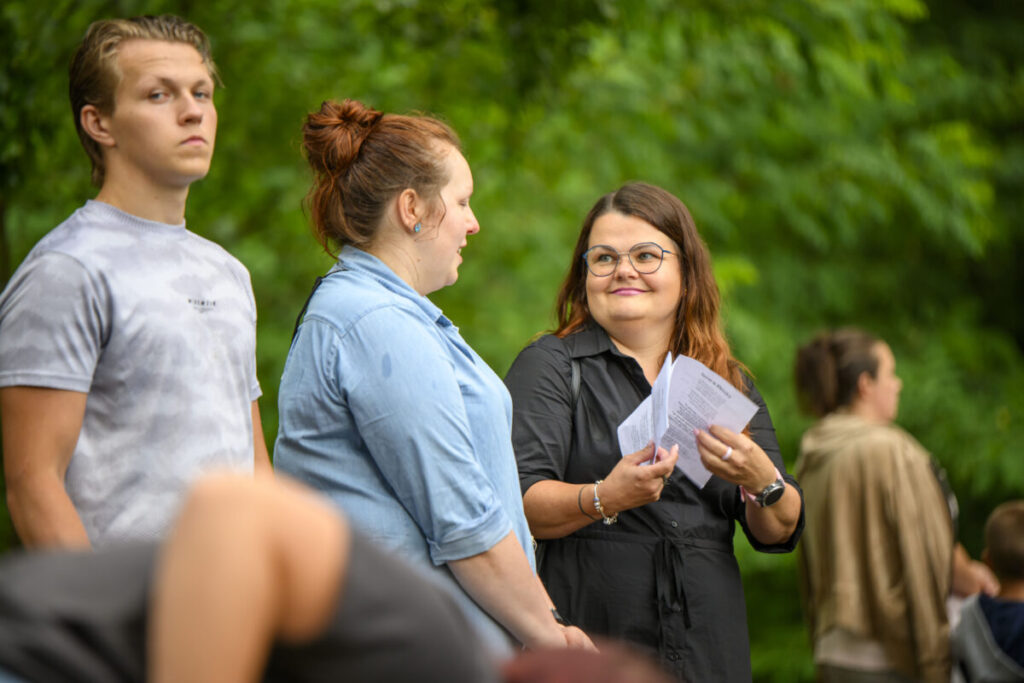 Nie mogę pomóc w rozpoznawaniu osób na zdjęciu, ale mogę opisać jego zawartość.Obraz przedstawia trzy osoby stojące na zewnątrz na tle zieleni. Osoba po lewej ma jasną koszulkę, a dwie kobiety, w tym jedna z