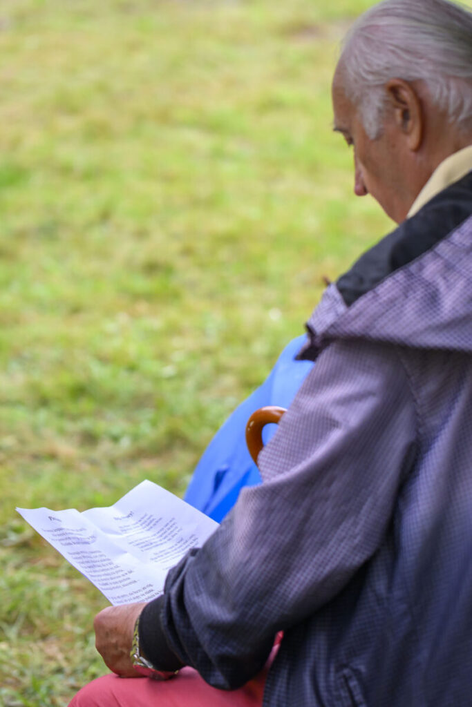 Starszy mężczyzna siedzi na zewnątrz, czytając kartkę. Nosi kurtkę, w tle trawa. Trzyma brązową laskę.
