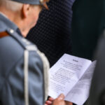 Starszy mężczyzna w mundurze czyta tekst z kartki. Mundur jest szary, tło rozmyte, wskazuje na zgromadzenie. Kartka zawiera wiersz Piechota.