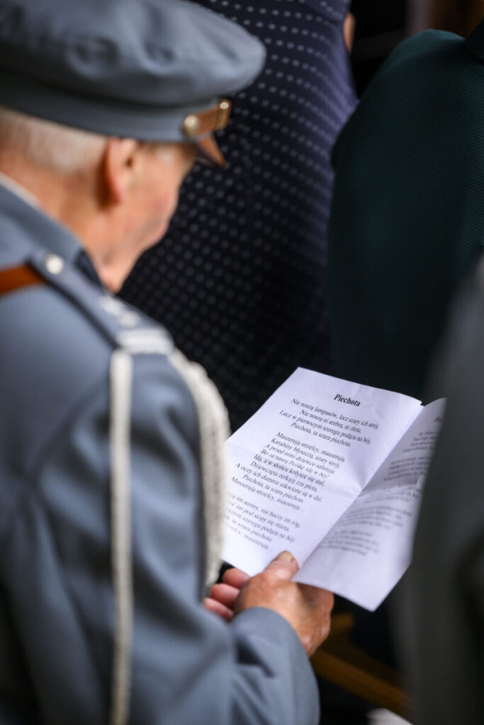 Starszy mężczyzna w mundurze czyta tekst z kartki. Mundur jest szary, tło rozmyte, wskazuje na zgromadzenie. Kartka zawiera wiersz Piechota.