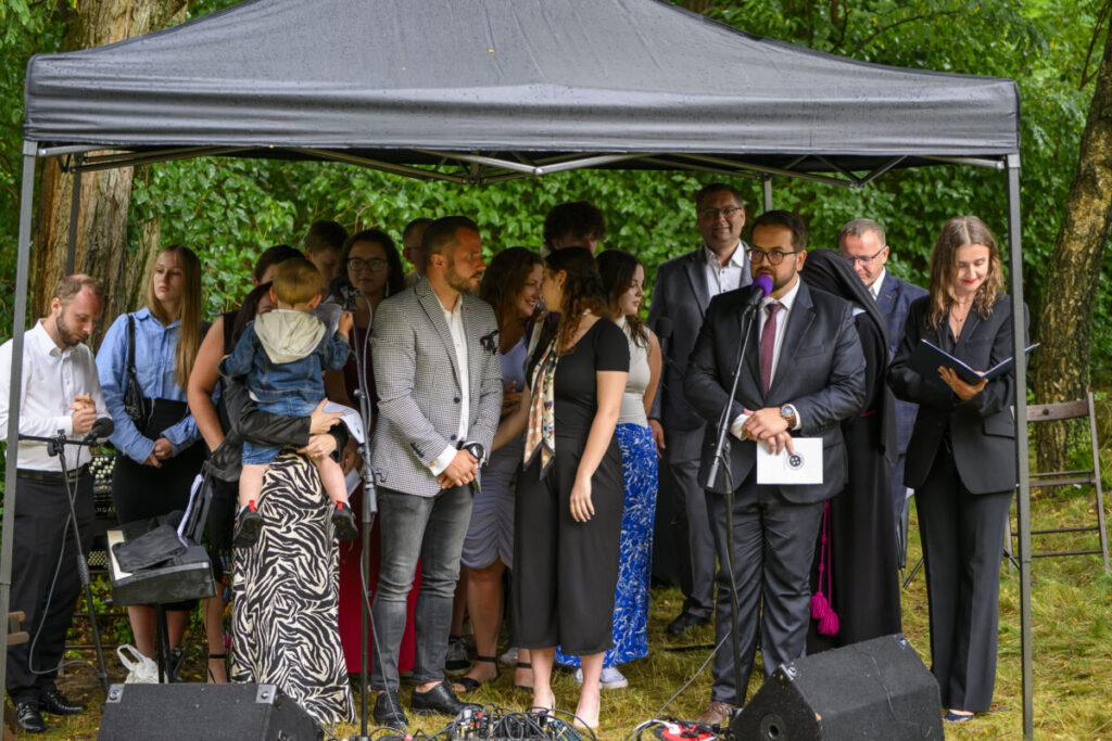 Grupa ludzi pod ciemnym namiotem, w centrum mężczyzna przy mikrofonie. W tle zielone krzewy, ziemia jest wilgotna. Atmosfera jest formalna.