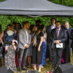 Grupa ludzi pod ciemnym namiotem, w centrum mężczyzna przy mikrofonie. W tle zielone krzewy, ziemia jest wilgotna. Atmosfera jest formalna.