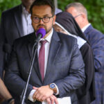 Mężczyzna w okularach, garniturze i z zegarkiem przemawia do mikrofonu z fioletową osłoną. W tle osoby w duchownych strojach i garniturach. Zielone liście tworzą rozmyte tło.