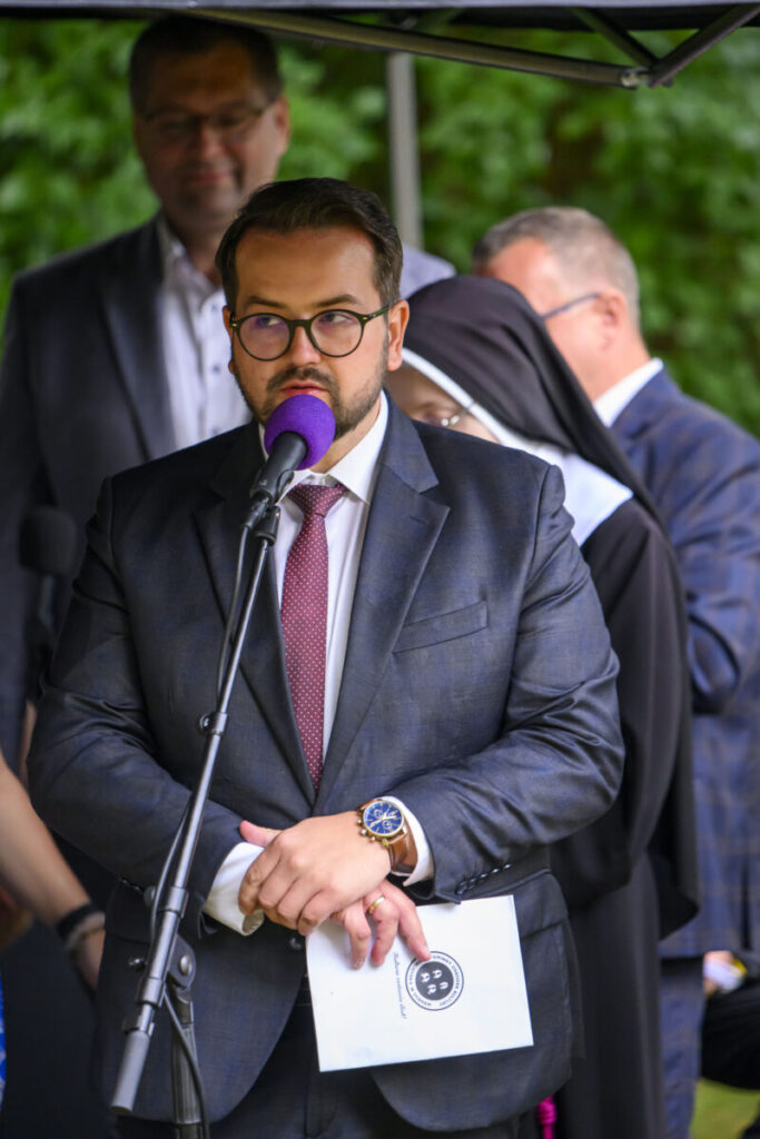 Mężczyzna w okularach, garniturze i z zegarkiem przemawia do mikrofonu z fioletową osłoną. W tle osoby w duchownych strojach i garniturach. Zielone liście tworzą rozmyte tło.