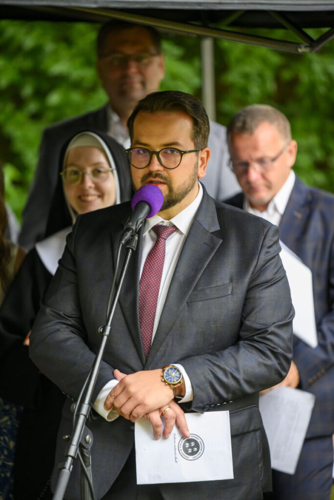 Mężczyzna w garniturze przemawia do mikrofonu, obok grupa ludzi. Tło jest zielone. Kobieta w habicie uśmiecha się.