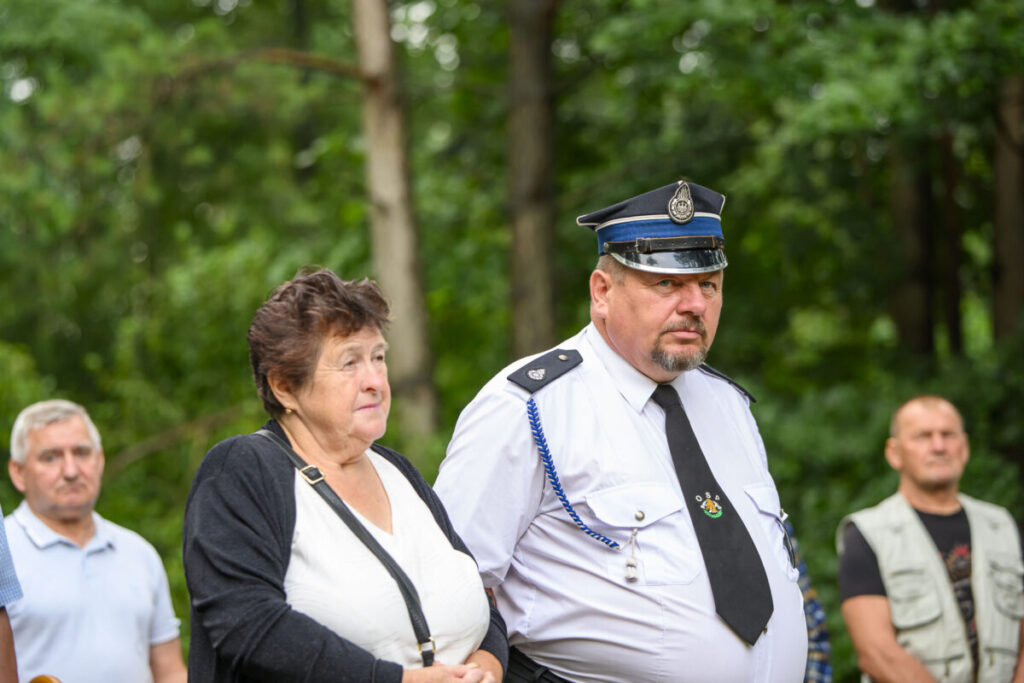 Na zdjęciu widać kobietę i mężczyznę w mundurze na tle zielonego lasu. Mężczyzna ma czapkę policyjną. Pozostali ludzie stoją w tle.