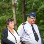 Na zdjęciu widać kobietę i mężczyznę w mundurze na tle zielonego lasu. Mężczyzna ma czapkę policyjną. Pozostali ludzie stoją w tle.