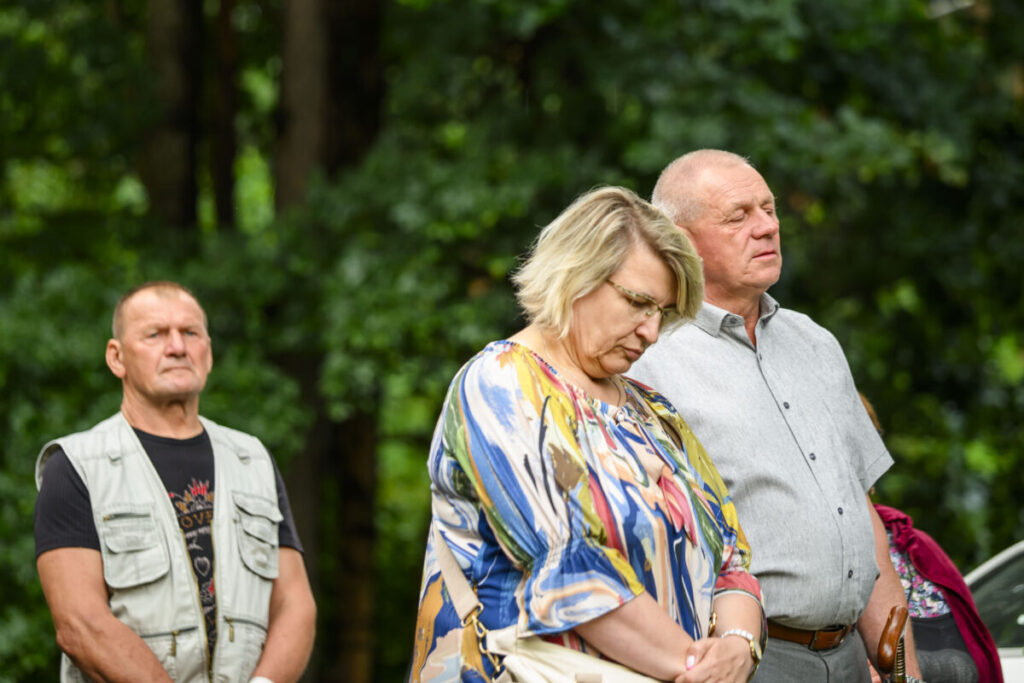 Trzy osoby stoją w lesie, patrząc w dół. Kobieta ma kolorową bluzkę, mężczyźni szare ubrania. Tło jest zielone i naturalne.